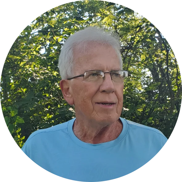 Portrait of author Gary Althaus: a white-haired man in glasses and a light blue t-shirt, standing in front of green trees.